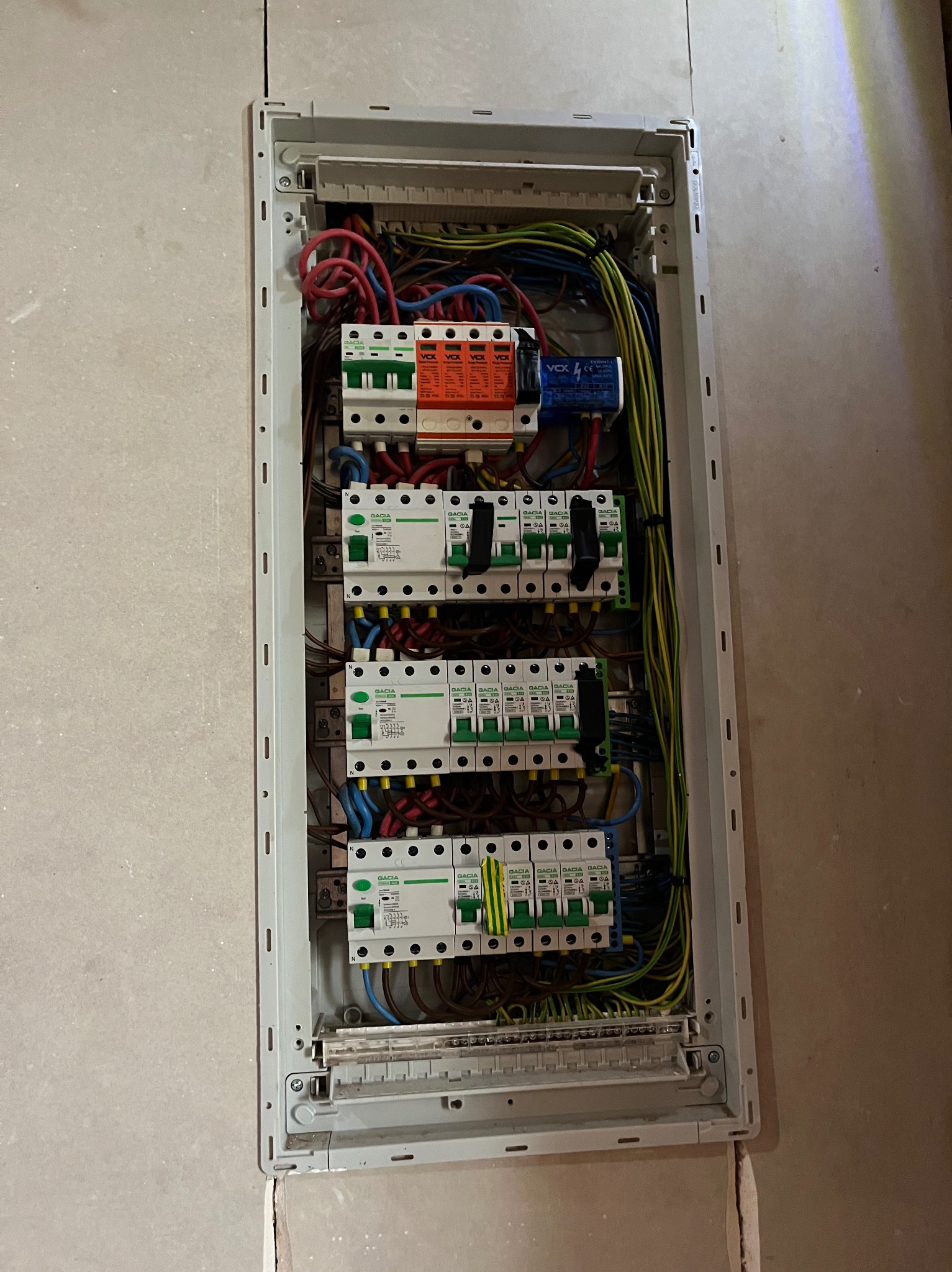 Szczegółowe ujęcie rozdzielnicy elektrycznej w jasnej obudowie, z widocznymi bezpiecznikami, przewodami w różnych kolorach i elementami zabezpieczeń.