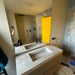 Usługi ogólnobudowlane Michał Wojciechowski - Nowoczesna, szara umywalka nablatowa z szafką w łazience podczas montażu. W tle widoczne narzędzia i fragment ściany z szarą mozaiką. W lustrze odbija się osoba robiąca zdjęcie.