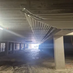 Równoległe rury instalacji pod stropem garażu, mocowane do sufitu za pomocą metalowych uchwytów. Widok w perspektywie, z kolumnami i odległym światłem.