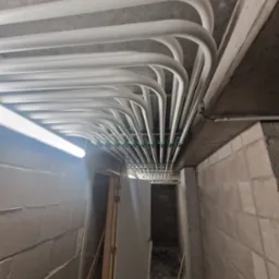 Białe rury instalacji podwieszane pod betonowym sufitem w piwnicy, z widocznym oświetleniem i fragmentem ściany z betonu komórkowego.