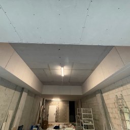 Usługi ogólnobudowlane Michał Wojciechowski - Sufit podwieszany z płyt gipsowo-kartonowych, widok z dołu na konstrukcję nośną i oświetlenie w surowym pomieszczeniu. Widoczne narzędzia i drabina.