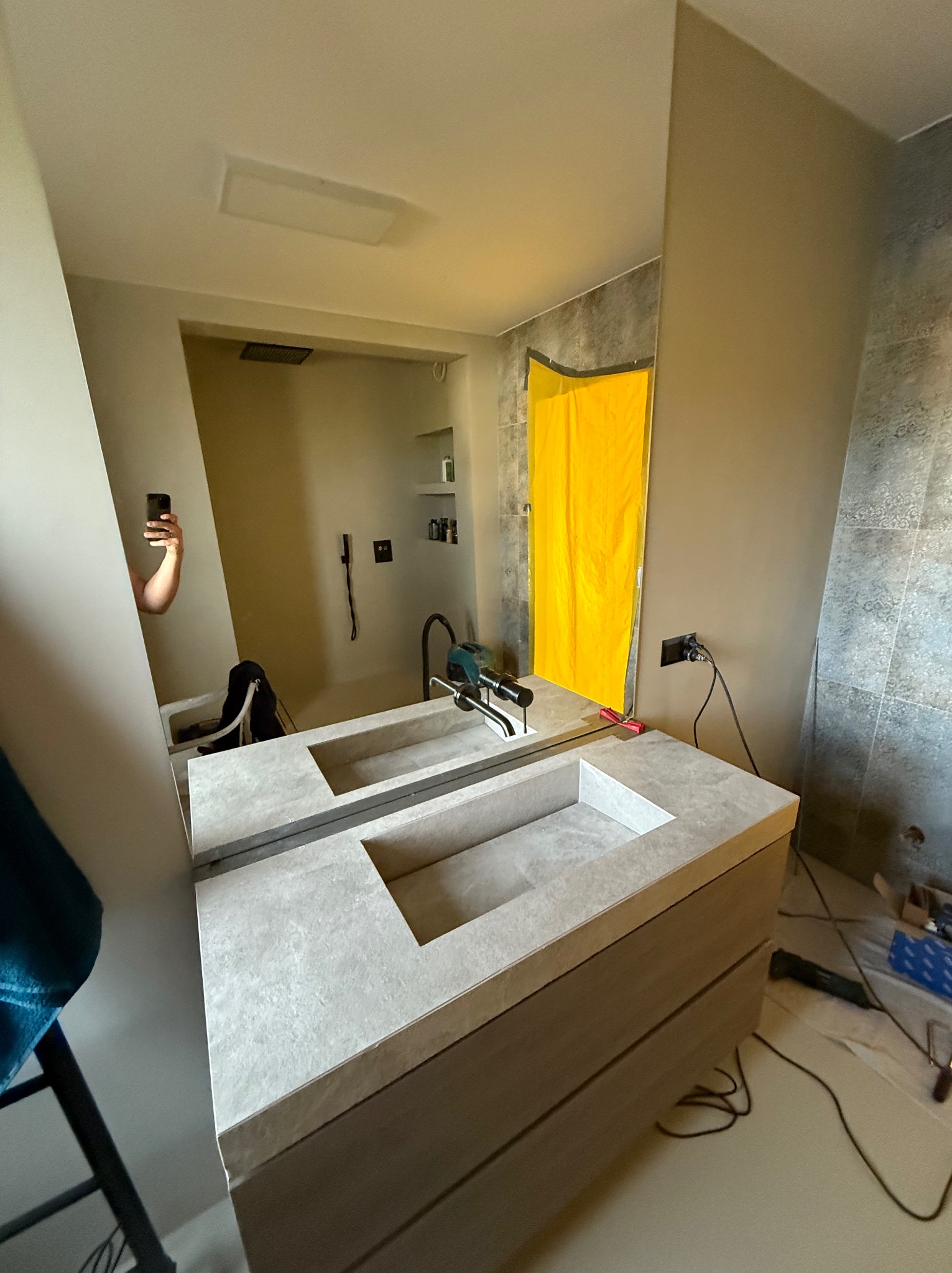 Nowoczesna, szara umywalka nablatowa z szafką w łazience podczas montażu. W tle widoczne narzędzia i fragment ściany z szarą mozaiką. W lustrze odbija się osoba robiąca zdjęcie.