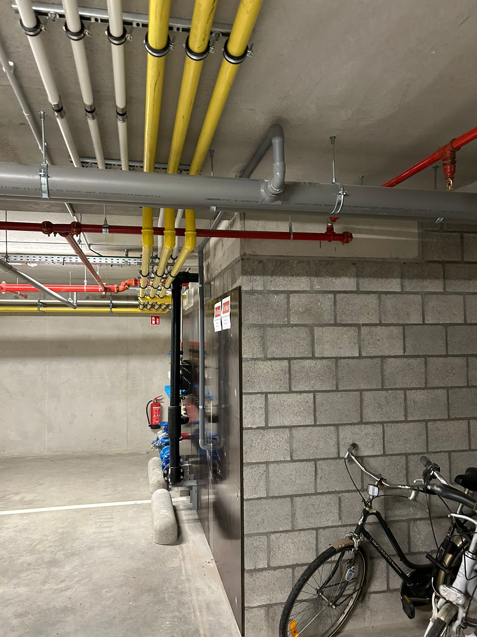 Różnokolorowe rury instalacyjne pod sufitem w Mogilnie. Widoczne rury białe, żółte, szare i czerwone. Na ścianie widoczny rower i gaśnica.