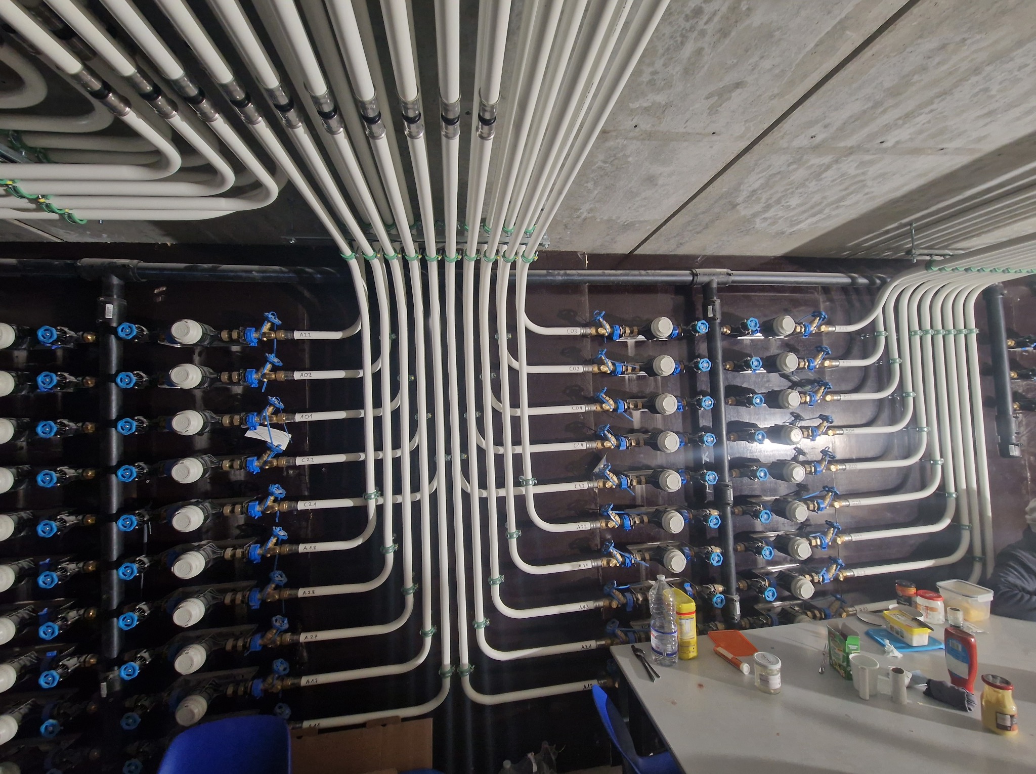 Rozbudowana instalacja hydrauliczna z białych rur i zaworów z niebieskimi pokrętłami, zamontowana na ciemnej ścianie, widoczna część sufitu i stół roboczy.
