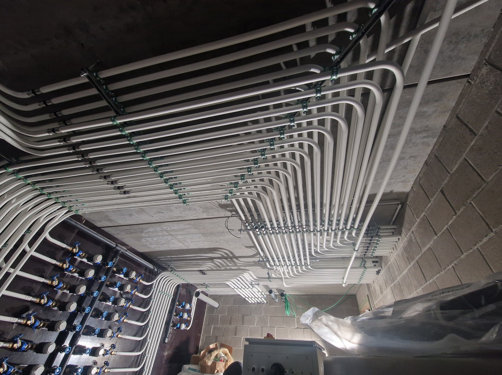 Równolegle ułożone białe rury instalacji wodnej pod stropem, mocowane zielonymi uchwytami, z widoczną ścianą z szarego betonu komórkowego.