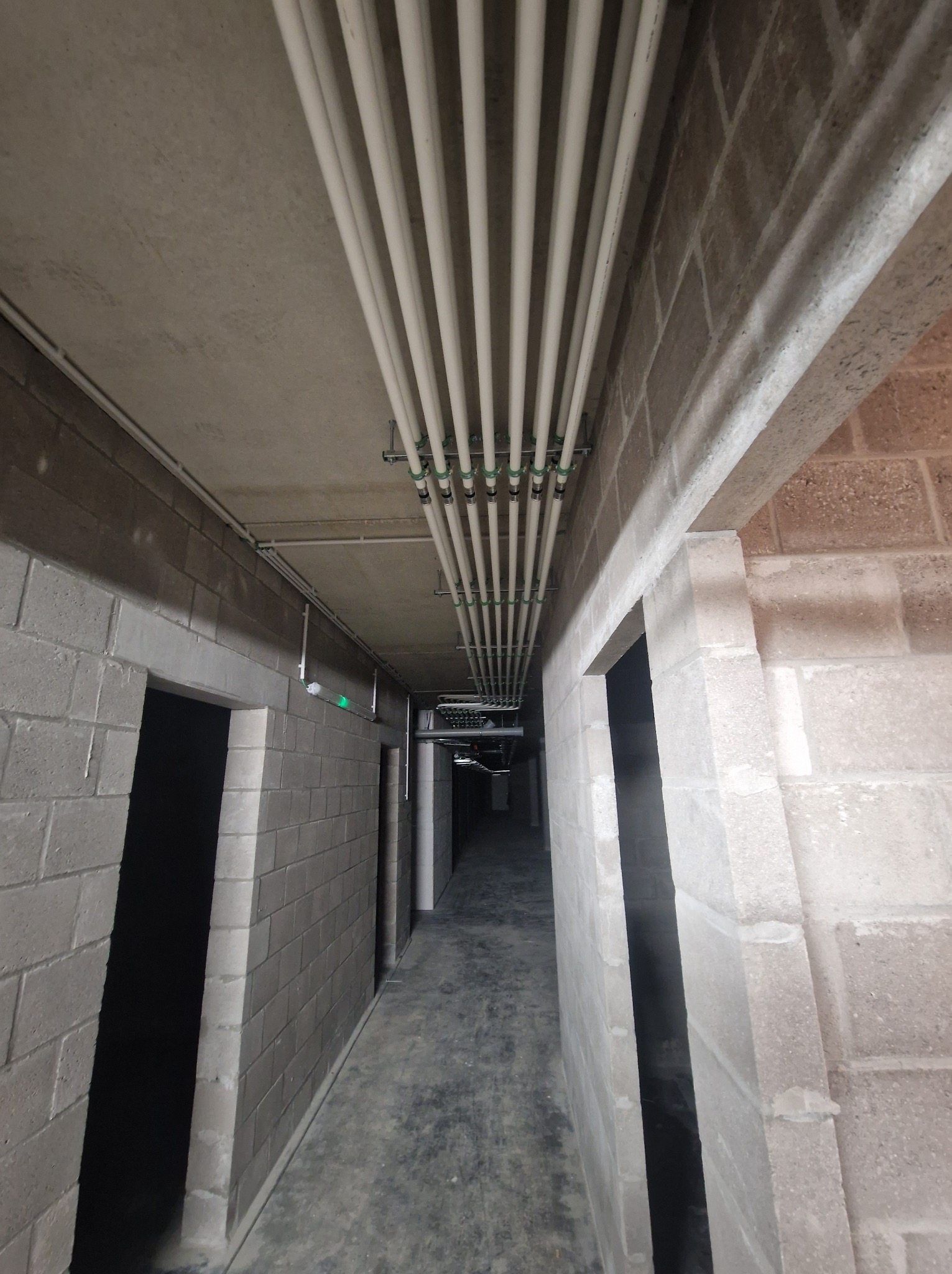 Wnętrze budynku w trakcie instalacji rur pod sufitem. Widoczne szare ściany z bloków i korytarz w perspektywie. Rury zamontowane równolegle.