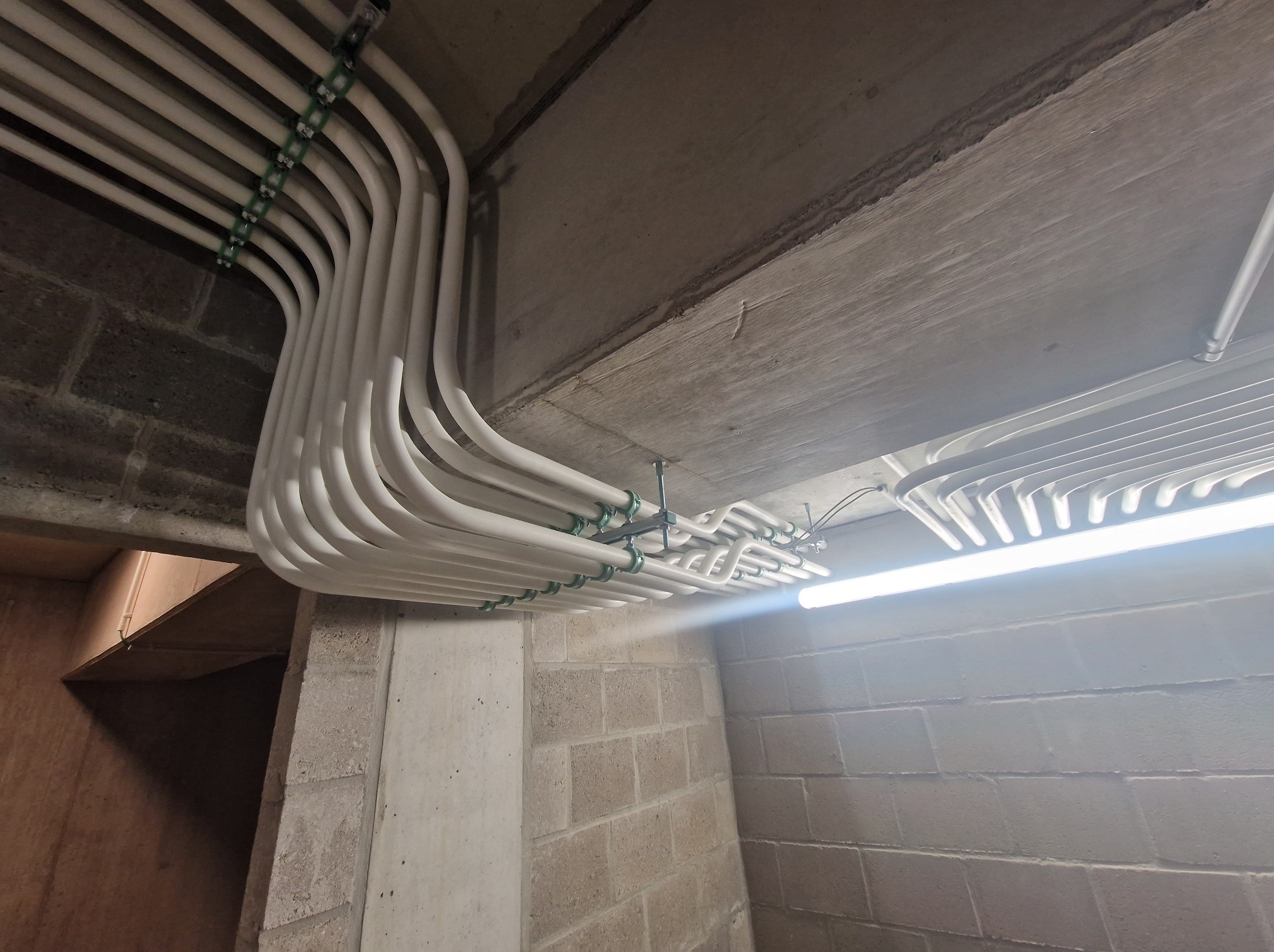 Białe rury instalacyjne równolegle ułożone pod betonowym sufitem, mocowane zielonymi uchwytami, oświetlone lampą jarzeniową, na tle ściany z szarego betonu komórkowego.