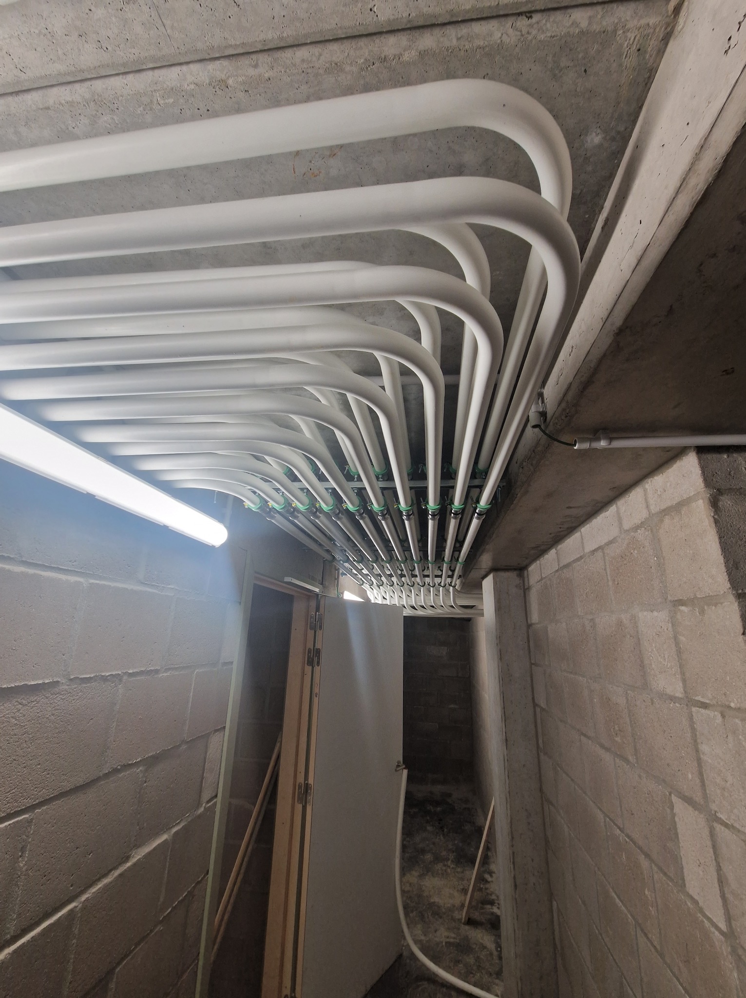 Białe rury instalacji podwieszane pod betonowym sufitem w piwnicy, z widocznym oświetleniem i fragmentem ściany z betonu komórkowego.