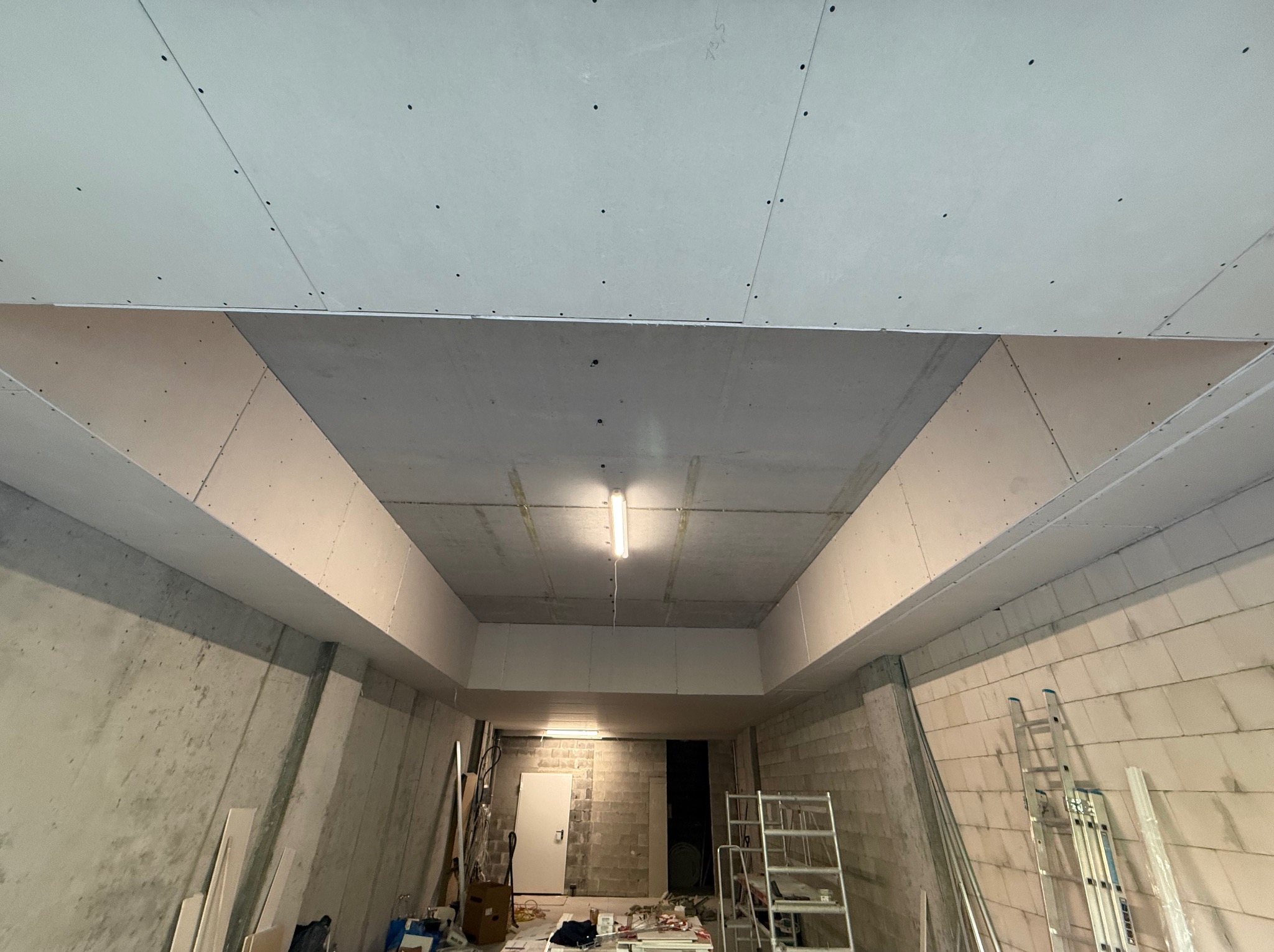 Sufit podwieszany z płyt gipsowo-kartonowych, widok z dołu na konstrukcję nośną i oświetlenie w surowym pomieszczeniu. Widoczne narzędzia i drabina.