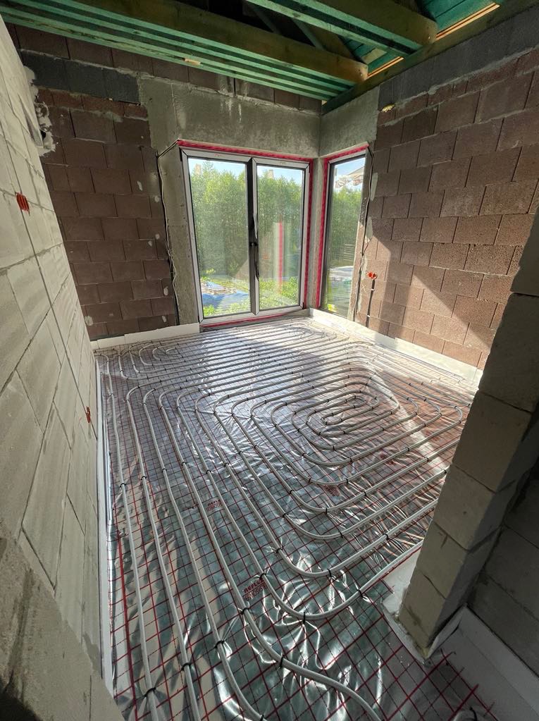 Nowoczesna instalacja ogrzewania podłogowego w surowym wnętrzu z oknem balkonowym. Białe rury ułożone na folii refleksyjnej, ściany z cegły i betonu.