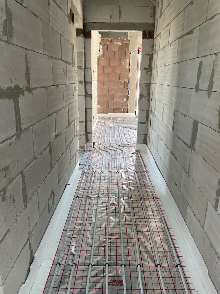 Układanie instalacji ogrzewania podłogowego na folii refleksyjnej w wąskim korytarzu z surowymi ścianami z betonu komórkowego przed wylewką. Widok z góry.