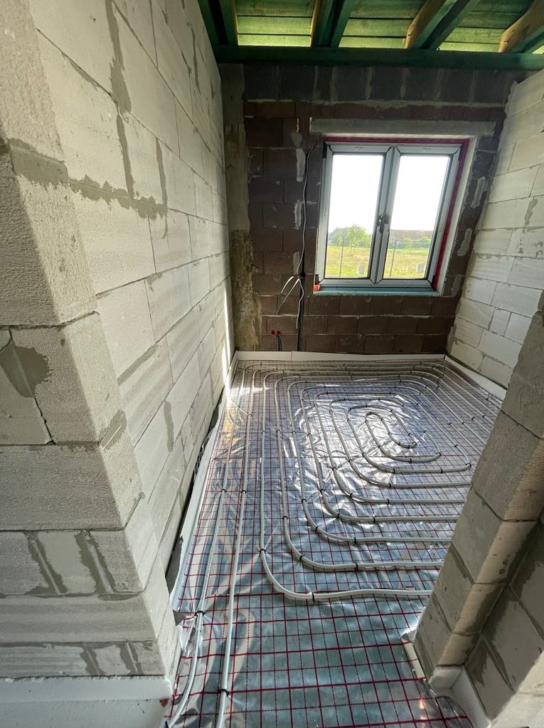 Białe rury ogrzewania podłogowego ułożone na folii z czerwoną siatką w surowym pomieszczeniu z oknem i ścianami z betonu komórkowego.