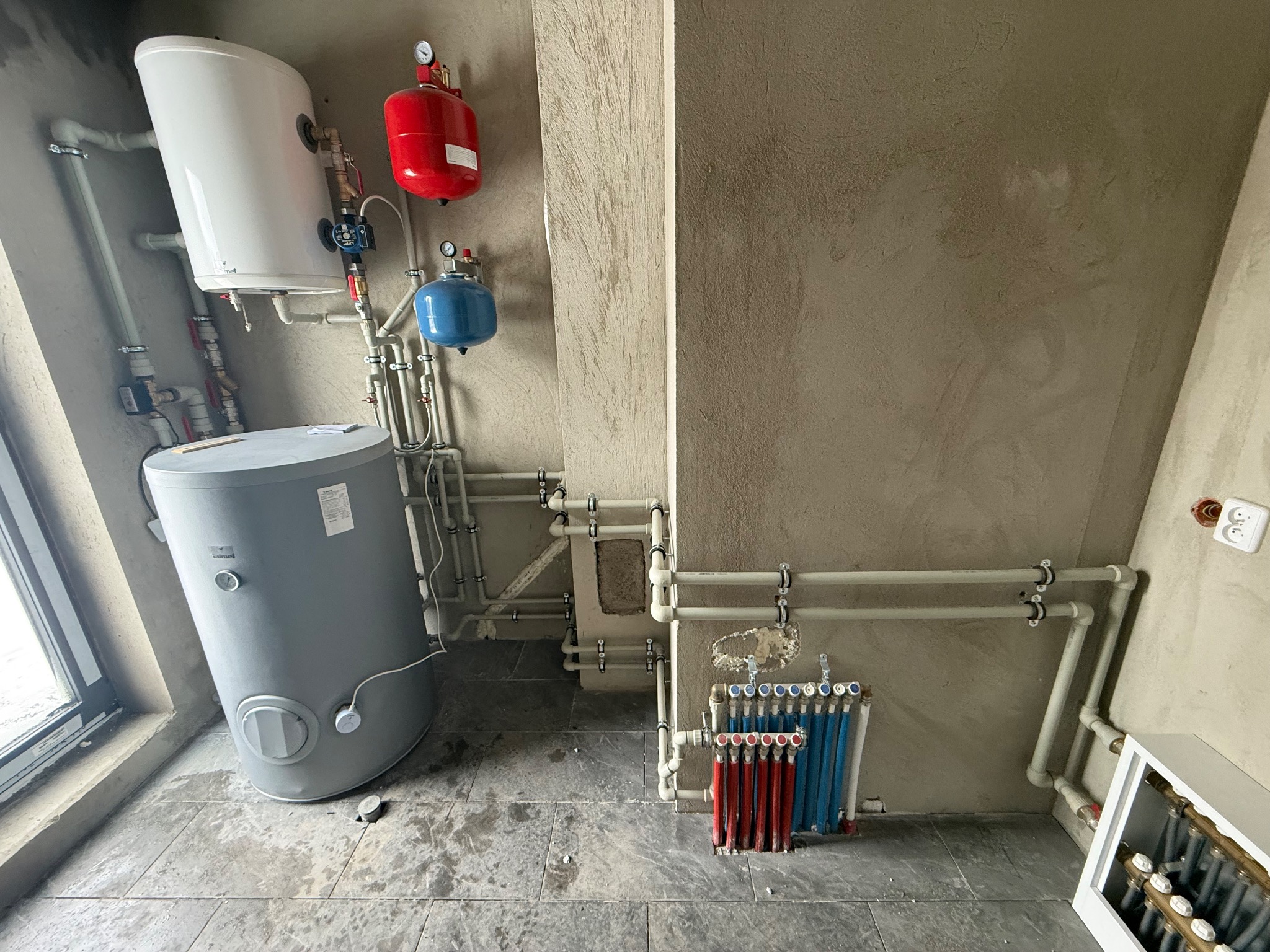 Kompleksowa instalacja CO z zbiornikami wyrównawczymi, grzejnikami i rozdzielaczem podłogówki. Rury prowadzone wzdłuż surowych ścian. Szare płytki podłogowe.