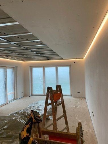 Wnętrze w trakcie prac wykończeniowych: widoczny sufit podwieszany z oświetleniem LED, drabina i zakryte folią okna.