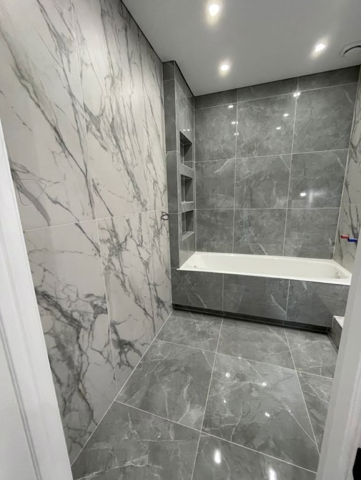 Wykończona łazienka z szarymi i białymi płytkami imitującymi marmur, zabudowana wanna i wnęki w ścianie