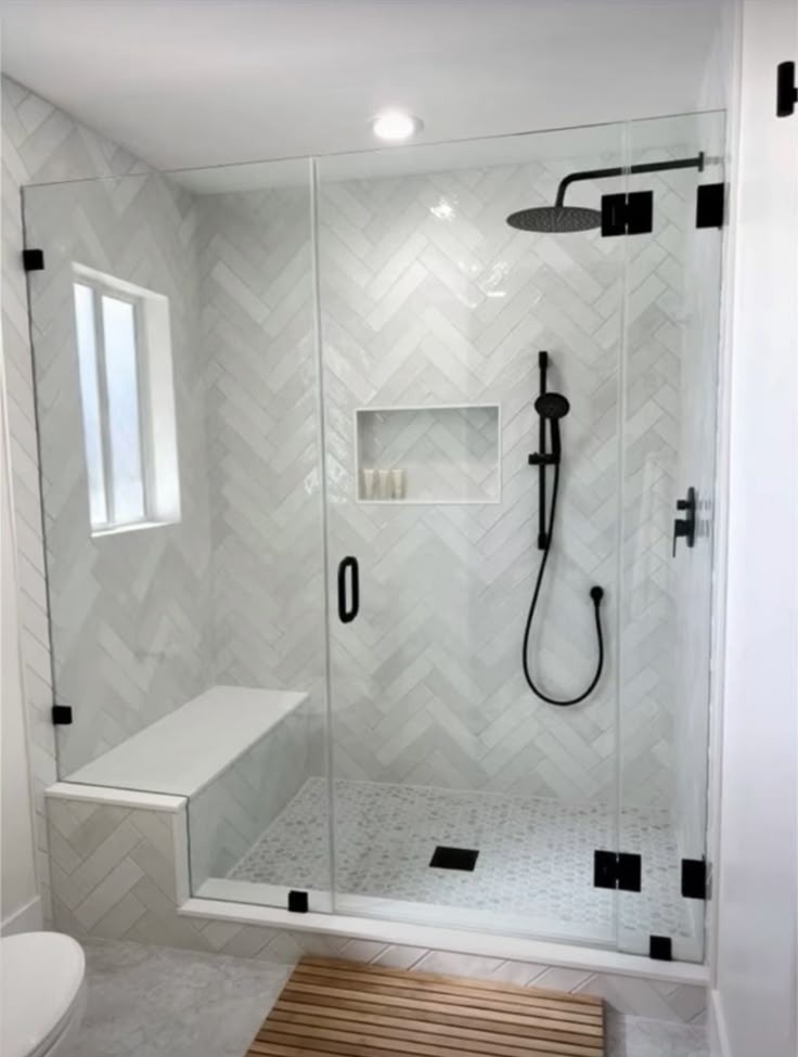 Nowoczesna kabina prysznicowa z białymi płytkami ułożonymi w jodełkę, czarną armaturą i szklanymi drzwiami, uzupełniona drewnianą matą podłogową.