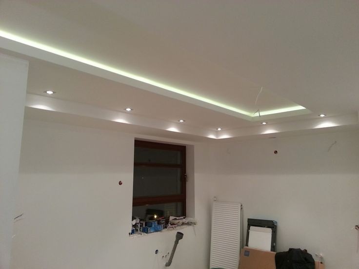 Wnętrze pokoju w trakcie prac wykończeniowych, widoczny podwieszany sufit z oświetleniem LED, okno z ciemną ramą, grzejnik, oraz drobne narzędzia i materiały budowlane na parapecie.