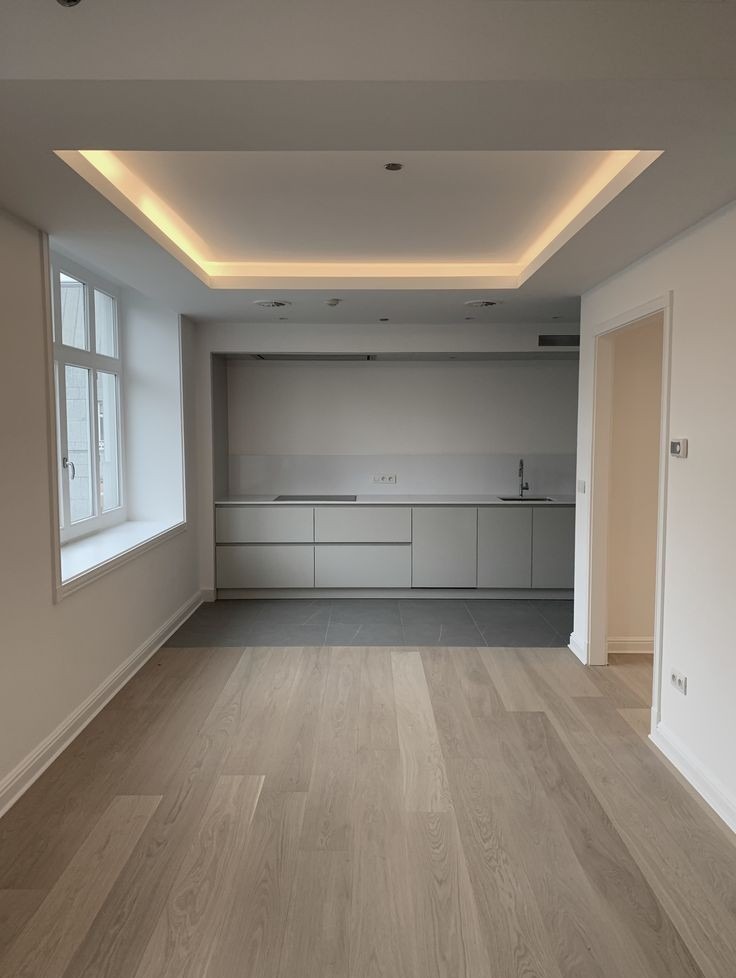 Nowoczesne wnętrze mieszkania po remoncie: parkiet w odcieniach szarości, białe ściany z listwami przypodłogowymi, minimalistyczna kuchnia w tle z szarymi frontami szafek i blatem, sufit...