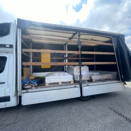 Marcin Ciastoń - Biały samochód dostawczy z otwartą plandeką, załadowany paletami z płytkami ceramicznymi lub panelami, zabezpieczonymi folią stretch.