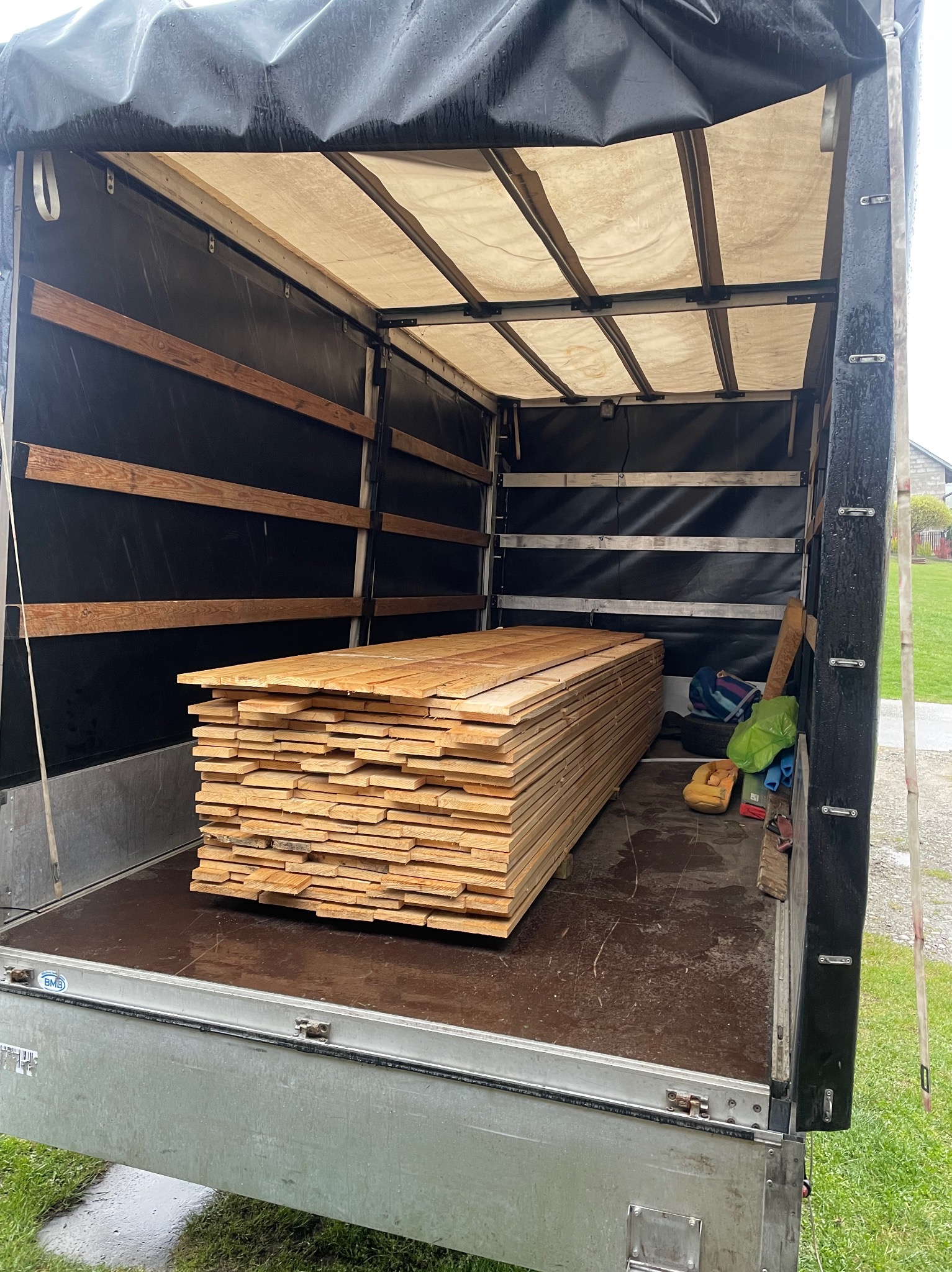 Stos desek z jasnego drewna ułożony w przestrzeni ładunkowej przyczepy z plandeką, widoczne zabezpieczenia ładunku i elementy konstrukcyjne przyczepy.