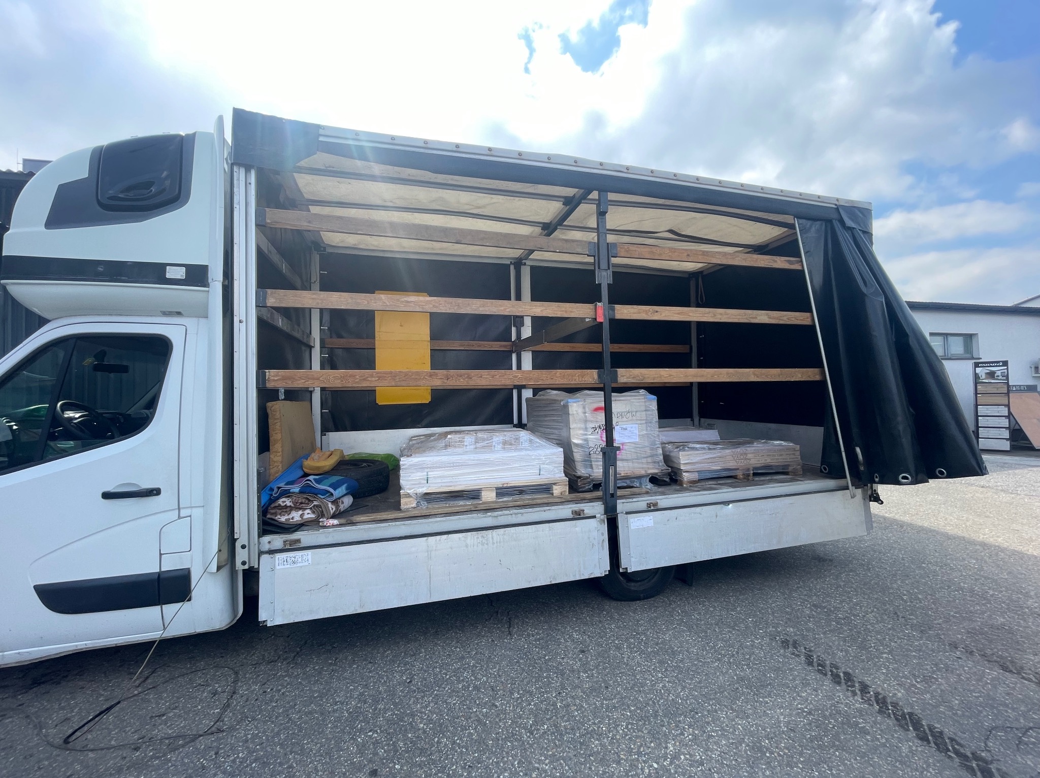 Biały samochód dostawczy z otwartą plandeką, załadowany paletami z płytkami ceramicznymi lub panelami, zabezpieczonymi folią stretch.