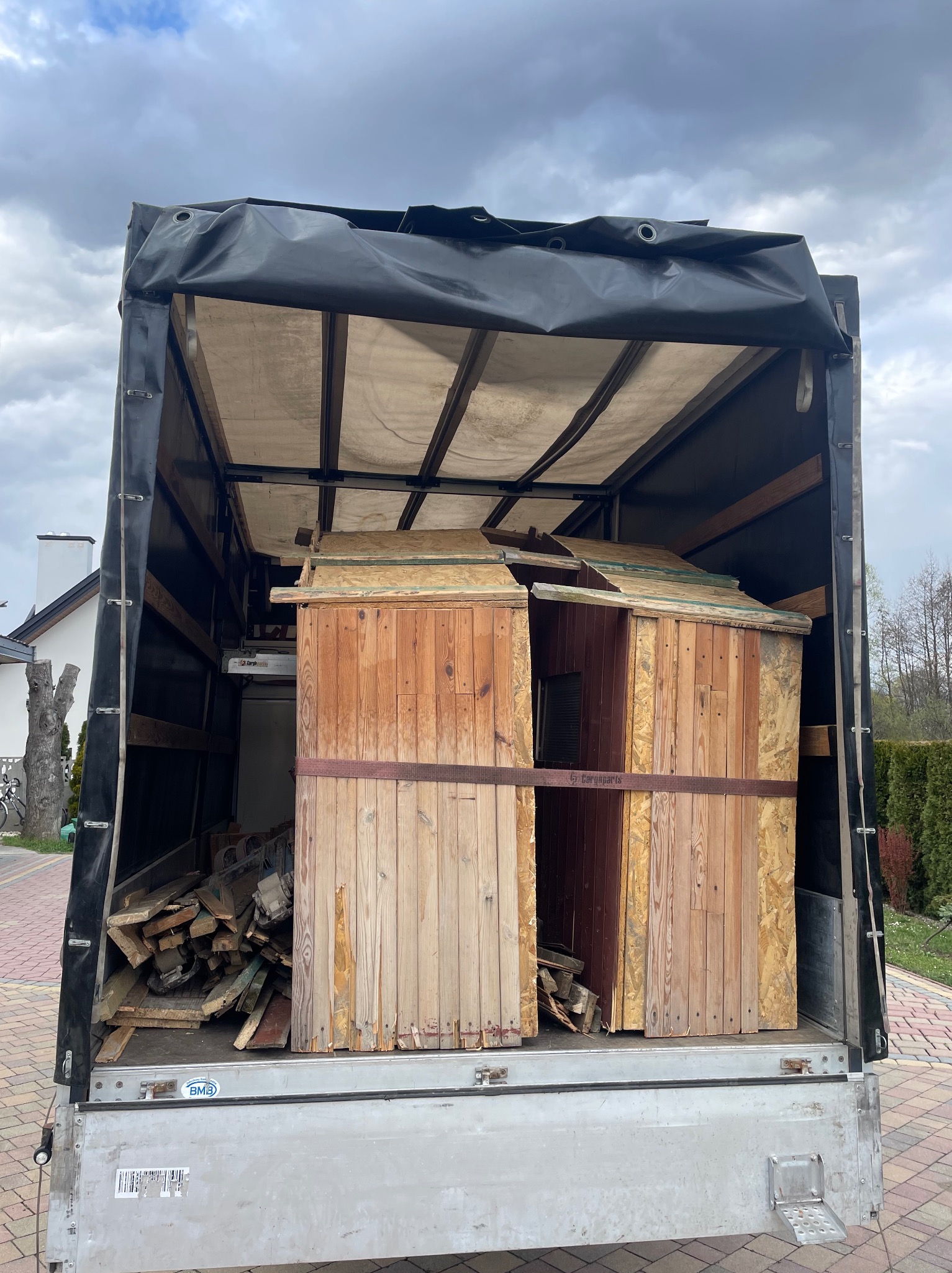 Otwarta paka ciężarówki z czarną plandeką, wypełniona elementami drewnianej konstrukcji, zabezpieczona brązowym pasem transportowym, obok leżą luźne deski.