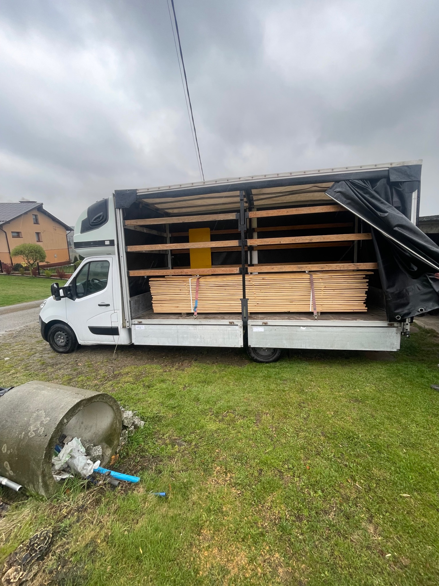 Biały bus z otwartą przestrzenią ładunkową, wypełniony drewnianymi deskami zabezpieczonymi pasami, z częściowo odsłoniętą plandeką, stojący na trawie obok betonowej rury wypełnionej śmieciami.