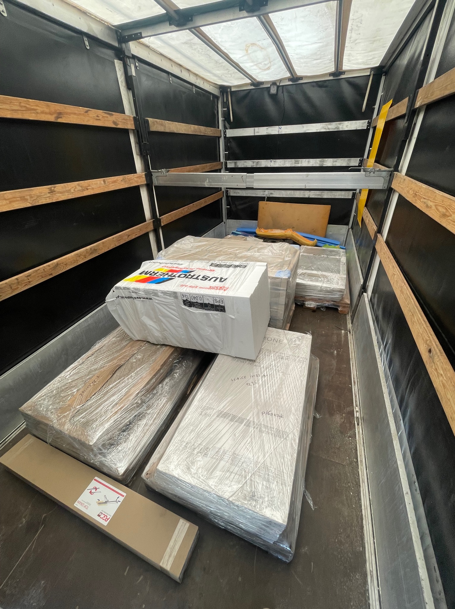 Wnętrze skrzyni ładunkowej wypełnionej paletami z zapakowanymi materiałami budowlanymi, w tym paczka styropianu Austrotherm, zabezpieczone folią stretch.