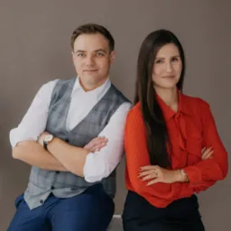 Portret dwojga prawników: mężczyzna w kraciastej kamizelce i kobieta w czerwonej bluzce, oboje z założonymi rękami, na neutralnym tle.