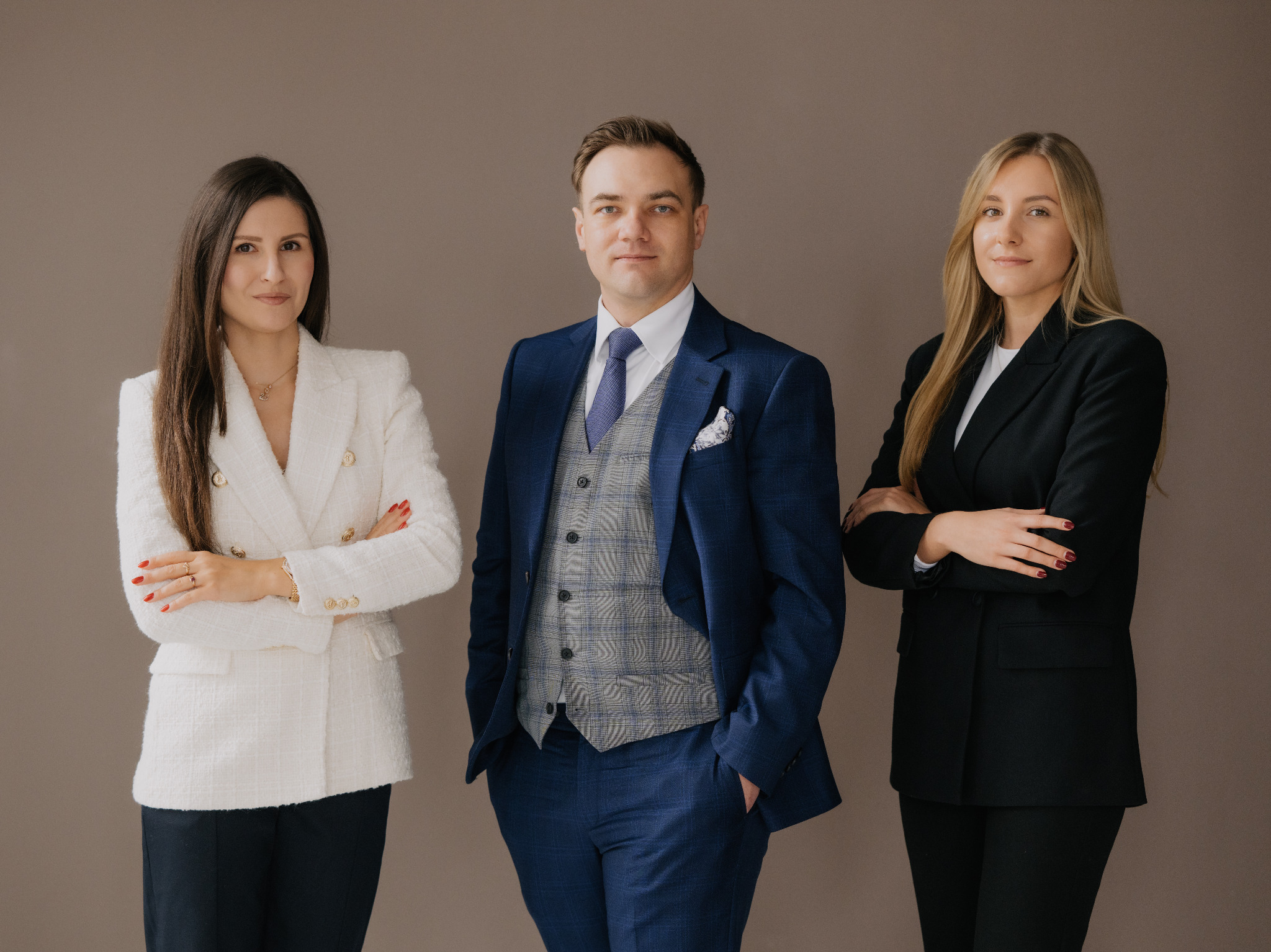 Profesjonalny portret grupowy: kobieta w białym żakiecie, mężczyzna w niebieskim garniturze i kamizelce w kratę, kobieta w czarnym żakiecie, wszyscy z założonymi rękami na neutralnym tle.