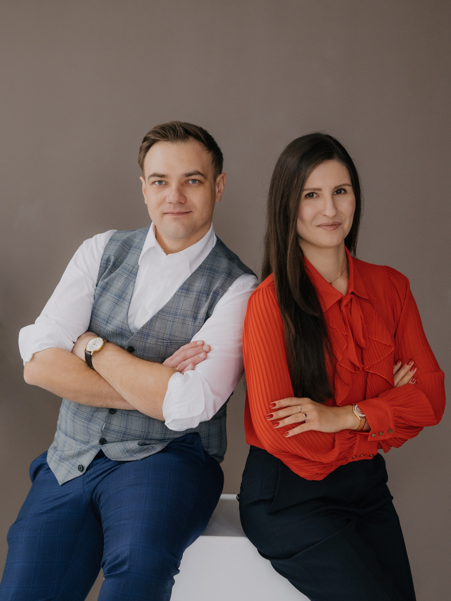 Portret dwojga prawników: mężczyzna w kraciastej kamizelce i kobieta w czerwonej bluzce, oboje z założonymi rękami, na neutralnym tle.