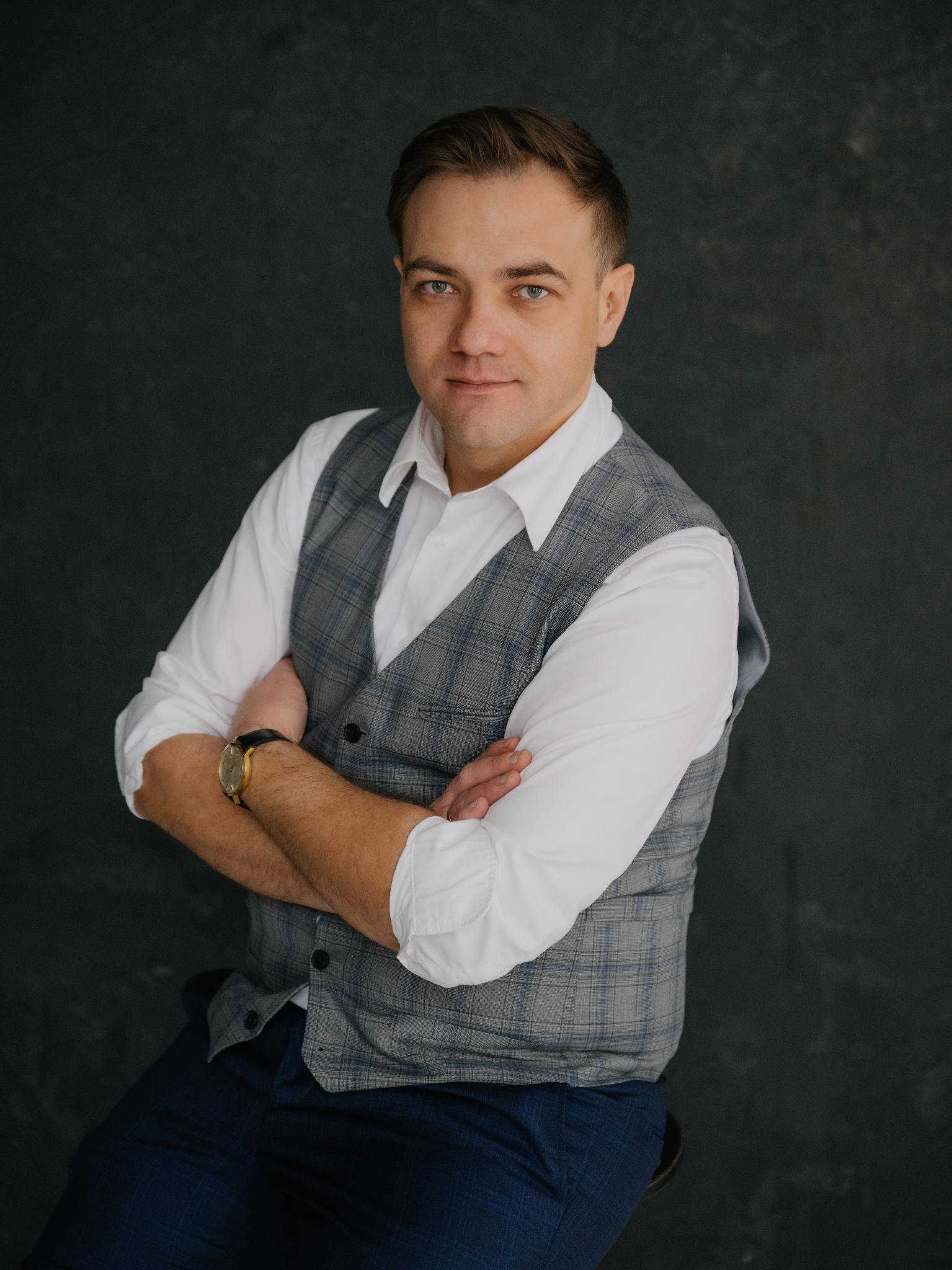 Portret mężczyzny w szarej kamizelce w kratę i białej koszuli, z założonymi rękami na tle ciemnej ściany, z widocznym złotym zegarkiem na ręku.