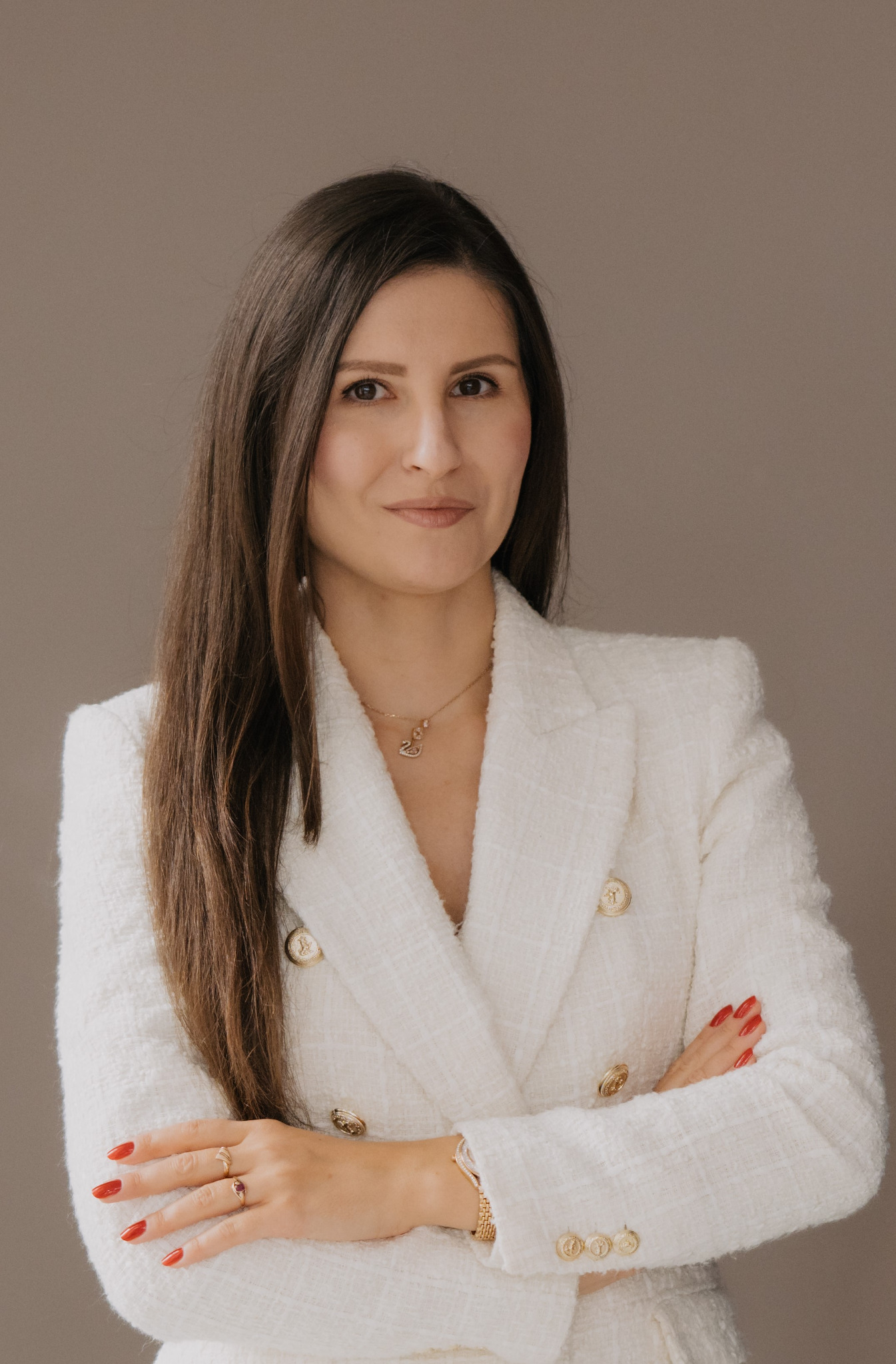 Portret kobiety w białym żakiecie z rękami założonymi na piersiach, z widoczną złotą biżuterią i czerwonym manicure na tle neutralnego beżu.