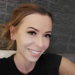 Portret uśmiechniętej kobiety z profesjonalnie wykonanym makijażem oczu, w tym z wyraźnie przedłużonymi rzęsami, na tle szarej ściany imitującej kamień.