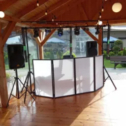 Scena plenerowa z nagłośnieniem i oświetleniem, biały parawan DJ-ski, drewniana podłoga i konstrukcja z żarówkami dekoracyjnymi.