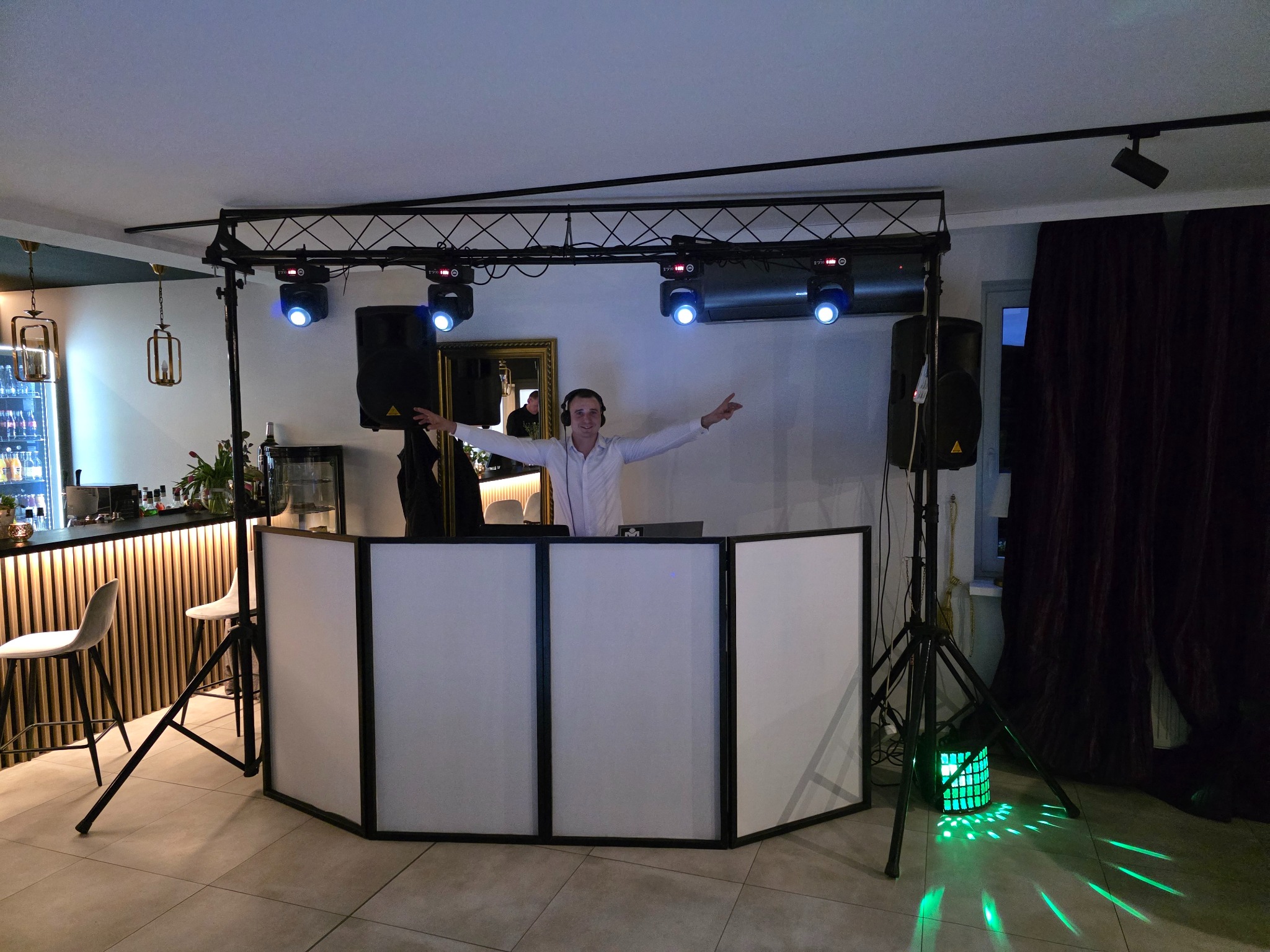 Uśmiechnięty DJ w białej koszuli, za białym panelem, z rozłożonymi rękami, na tle nagłośnienia i oświetlenia scenicznego z reflektorami i kolumnami, widoczny fragment baru.