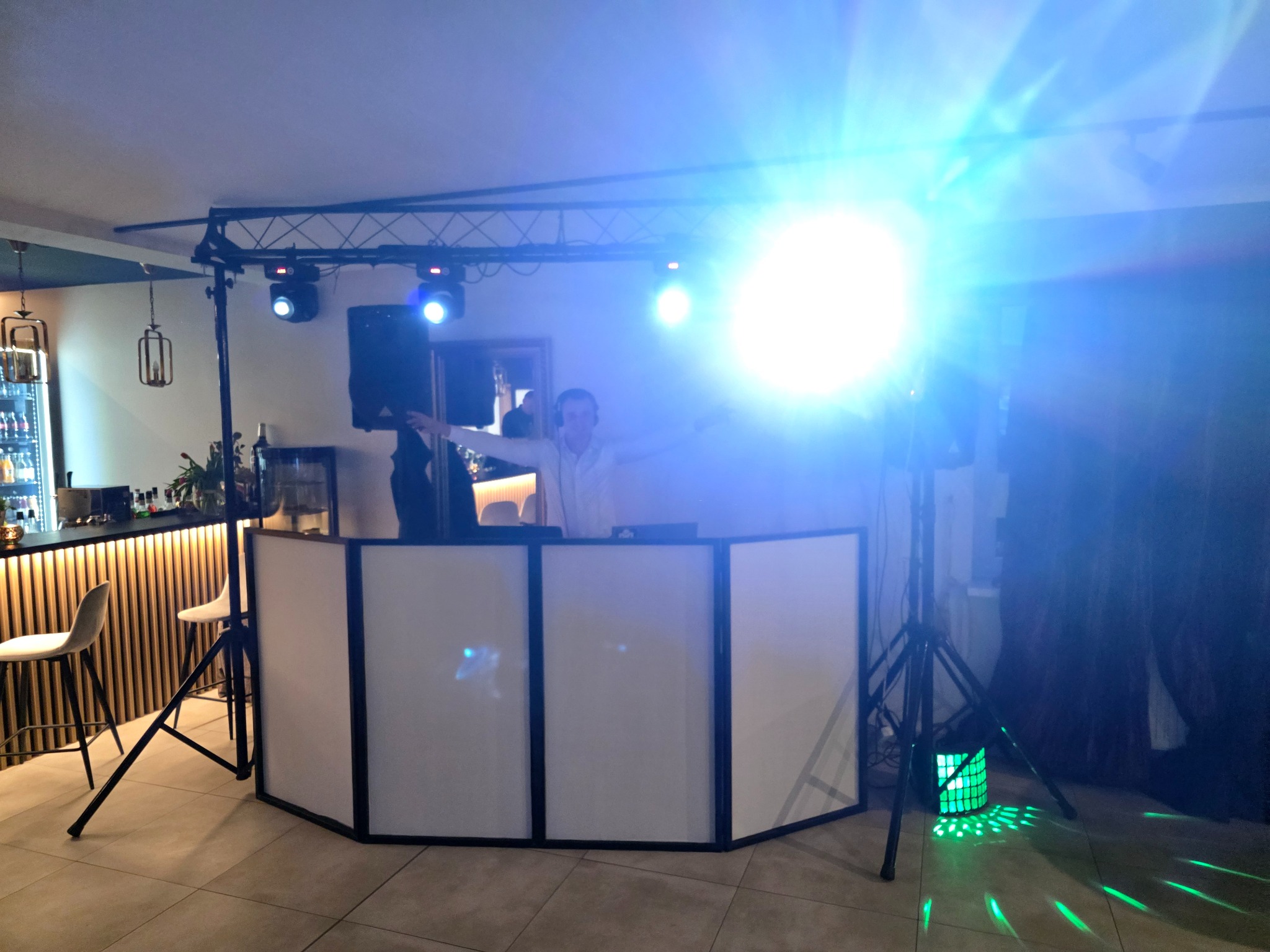 DJ za konsoletą z białym panelem, oświetlenie sceniczne z efektami świetlnymi, bar z alkoholem w tle