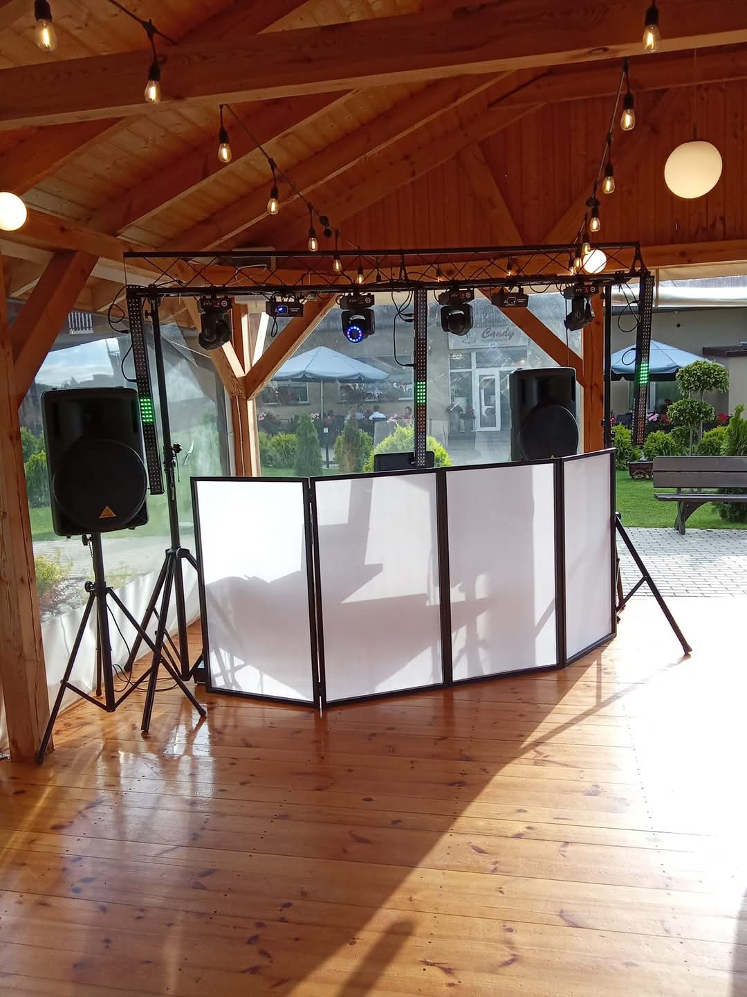 Scena plenerowa z nagłośnieniem i oświetleniem, biały parawan DJ-ski, drewniana podłoga i konstrukcja z żarówkami dekoracyjnymi.