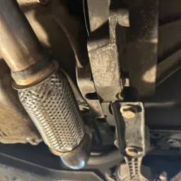 Widok od spodu na element układu wydechowego samochodu z metalowym łącznikiem elastycznym i mocowaniem, ukazujący detale konstrukcyjne pojazdu.