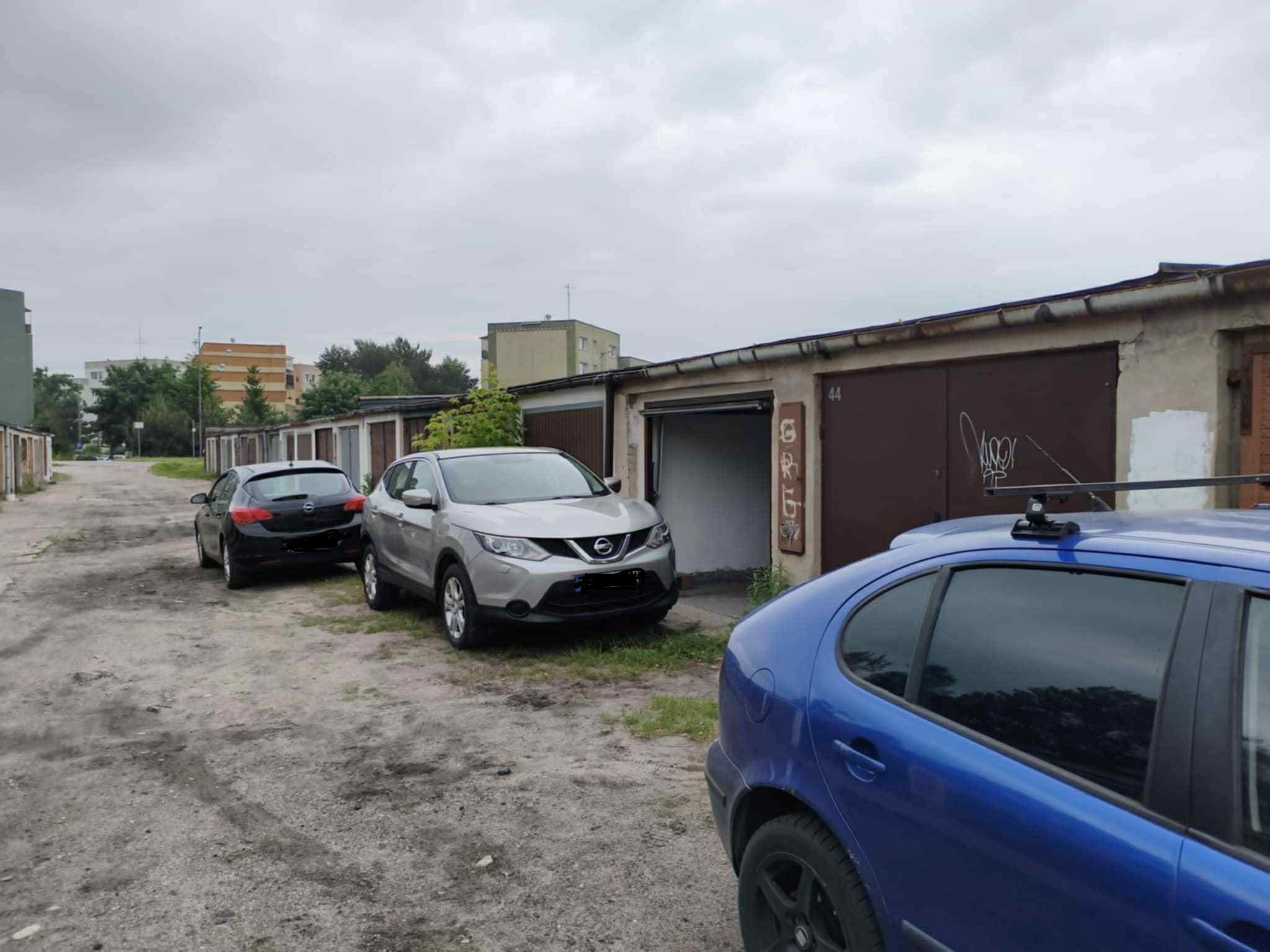 Samochody zaparkowane przed garażami z numerami, w pochmurny dzień. Widoczne budynki w tle i fragment nieba. Jeden z garaży ma otwarte drzwi.