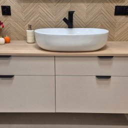 MRS SPÓŁKA Z OGRANICZONĄ ODPOWIEDZIALNOŚCIĄ - Nowoczesna szafka łazienkowa z blatem imitującym drewno, białą umywalką i czarną armaturą. Na blacie dekoracje: świeca, dozownik i złoty wazon z jesiennymi kwiatami.