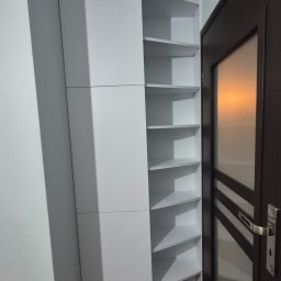 MRS SPÓŁKA Z OGRANICZONĄ ODPOWIEDZIALNOŚCIĄ - Biała szafa narożna z półkami, wkomponowana we wnękę obok drzwi. Minimalistyczny design, gładkie fronty, widoczne półki w kształcie trójkątów. Funkcjonalne rozwiązanie do przechowywania.