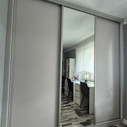 MRS SPÓŁKA Z OGRANICZONĄ ODPOWIEDZIALNOŚCIĄ - Jasnoszara szafa wnękowa z przesuwnymi drzwiami i lustrem, odbijająca pokój z biurkiem i krzesłami. Podłoga z paneli w odcieniach szarości i brązu.