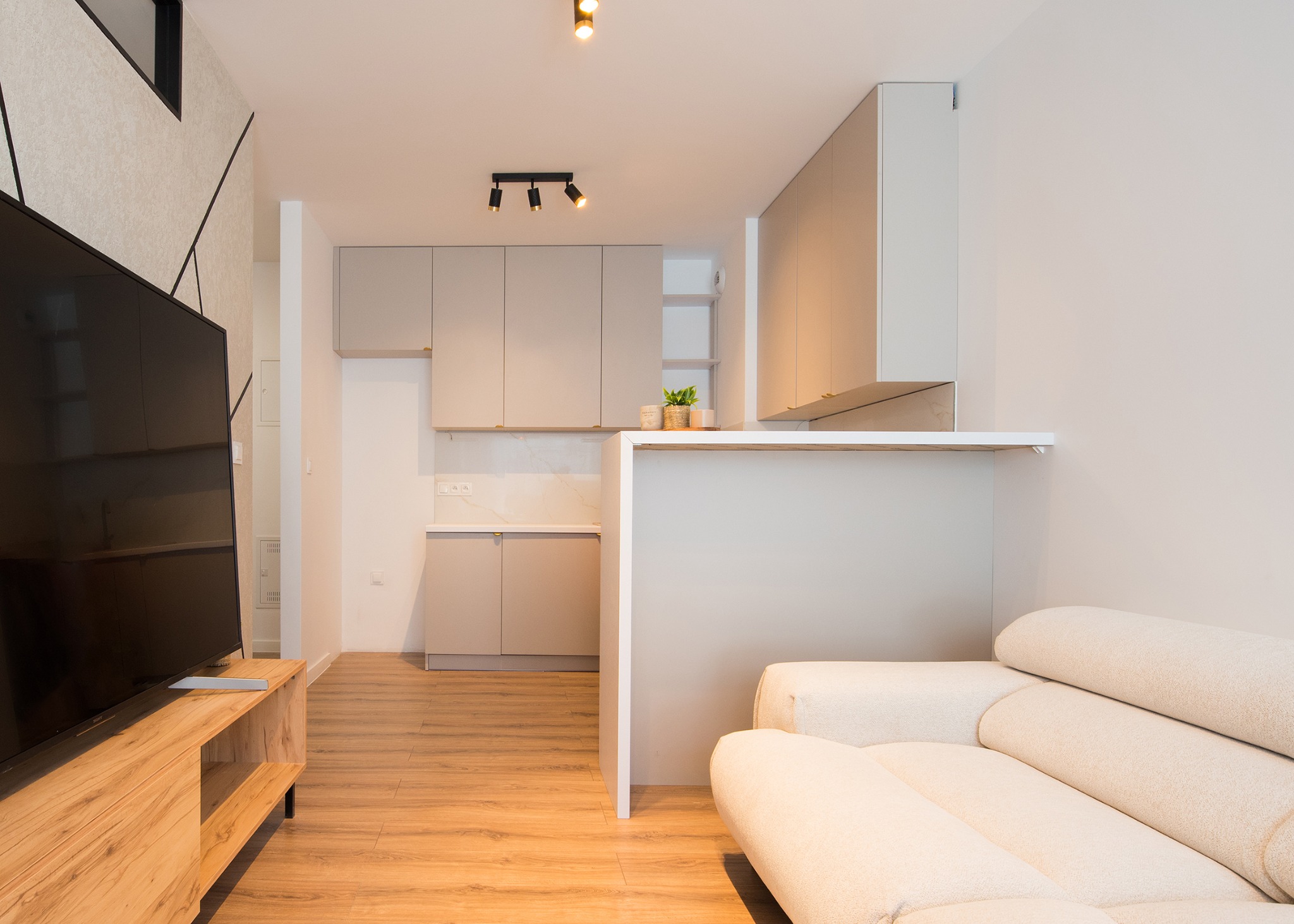 Nowoczesna kuchnia z szarymi frontami, częściowo otwarta na salon z drewnianą szafką RTV i beżową sofą. Widoczne oświetlenie punktowe i geometryczny wzór na ścianie.