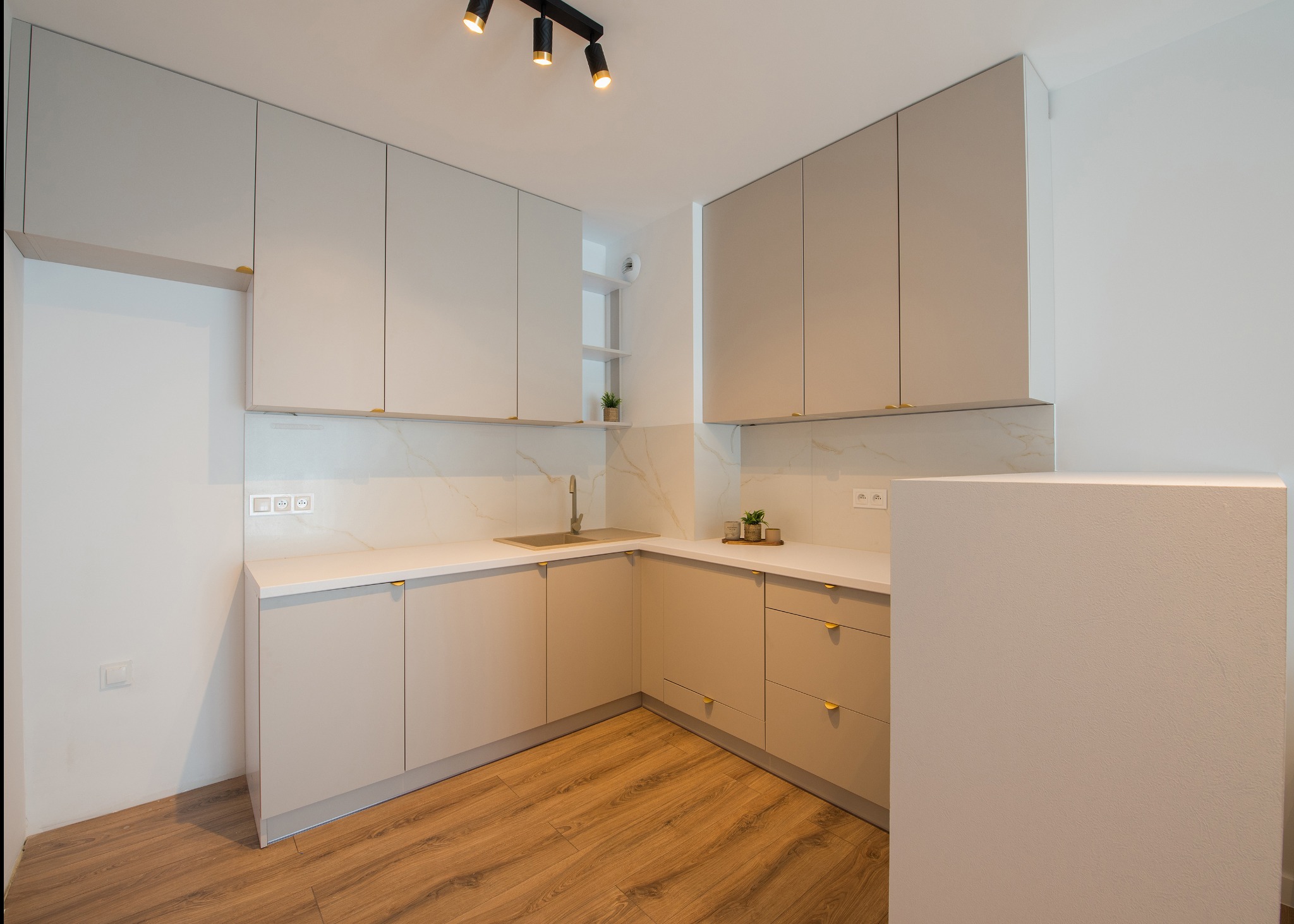 Jasnoszara zabudowa kuchenna z złotymi uchwytami, białym blatem i marmurowym backsplashiem, oświetlona reflektorami punktowymi