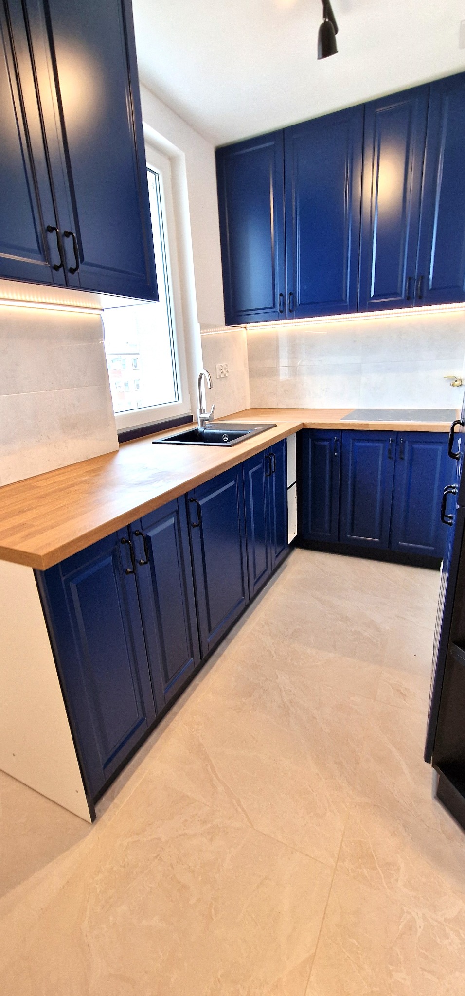 Nowoczesna, narożna kuchnia z granatowymi szafkami z frezowanymi frontami, drewnianym blatem i białymi płytkami nad blatem, oświetlona listwą LED.