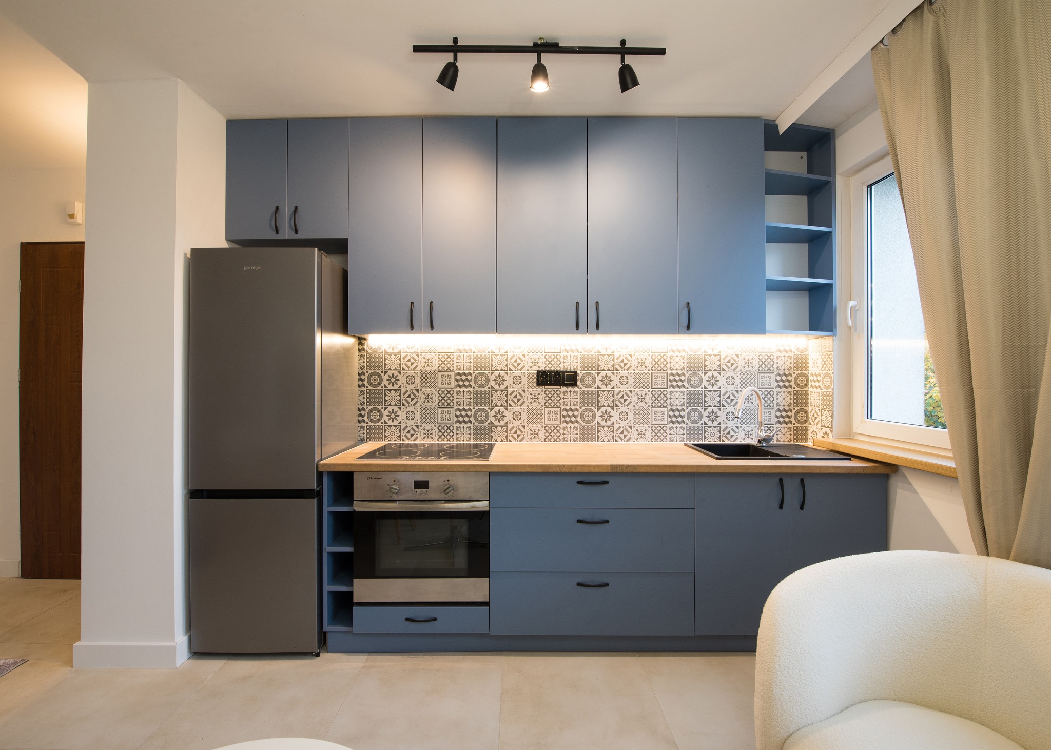 Nowoczesna kuchnia w odcieniach szarości i błękitu z drewnianym blatem, wzorzystymi płytkami nad blatem i oświetleniem punktowym, uzupełniona o lodówkę Gorenje i jasną zasłonę przy oknie.