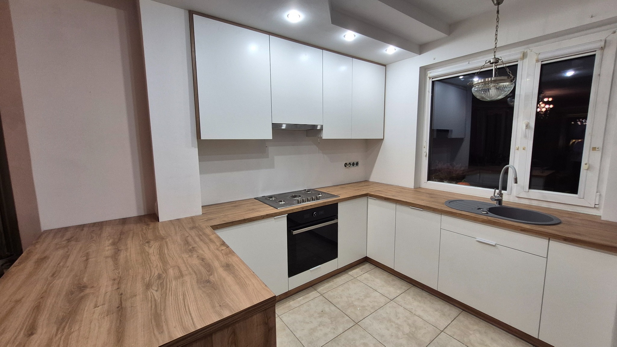 Nowoczesna kuchnia w kształcie litery U z białymi frontami i drewnianym blatem. Widoczna płyta grzewcza, piekarnik i okno z lampą. Minimalistyczny design.