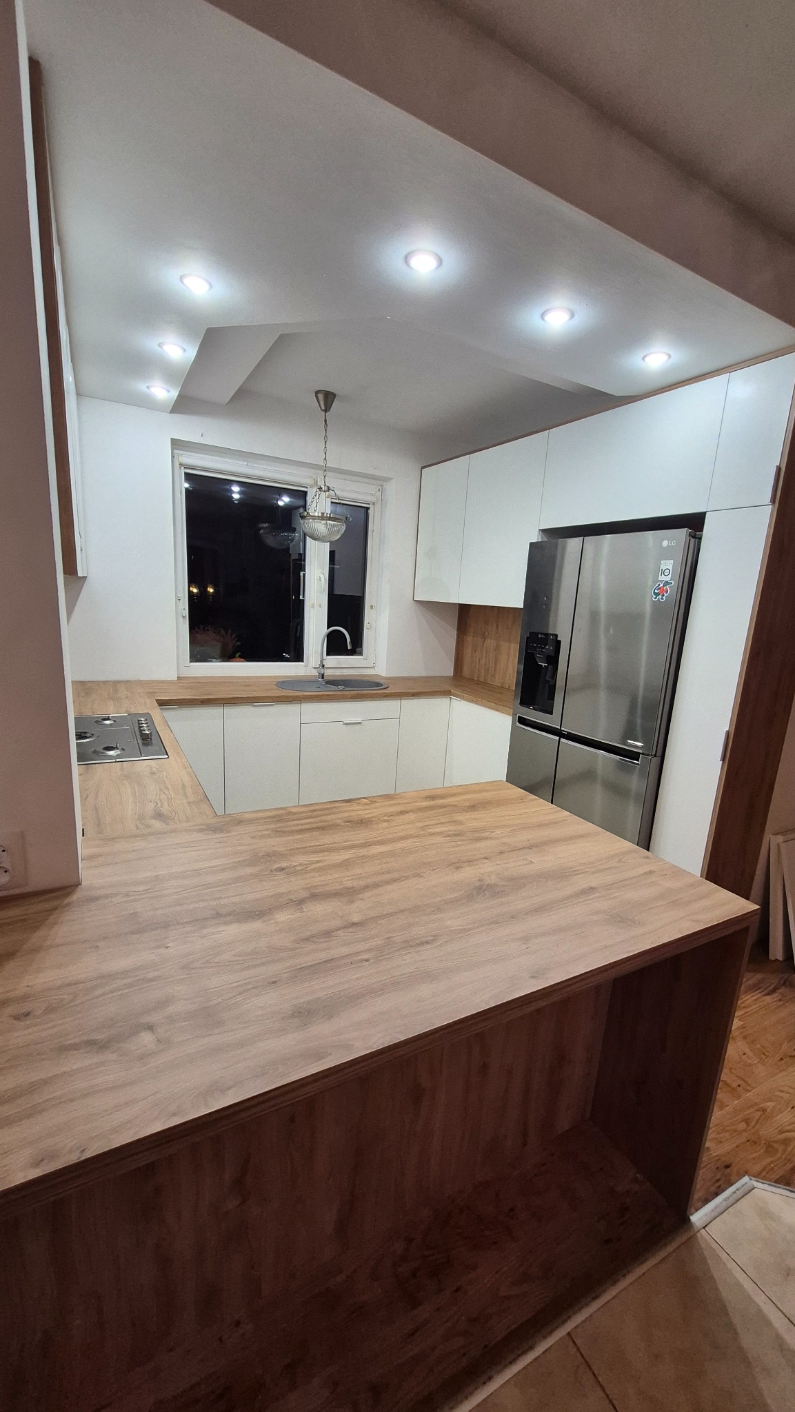 Nowoczesna kuchnia z białymi szafkami i drewnianym blatem, oświetlona punktowo, z wyspą kuchenną i srebrną lodówką. Widok na okno z dekoracyjną lampą.