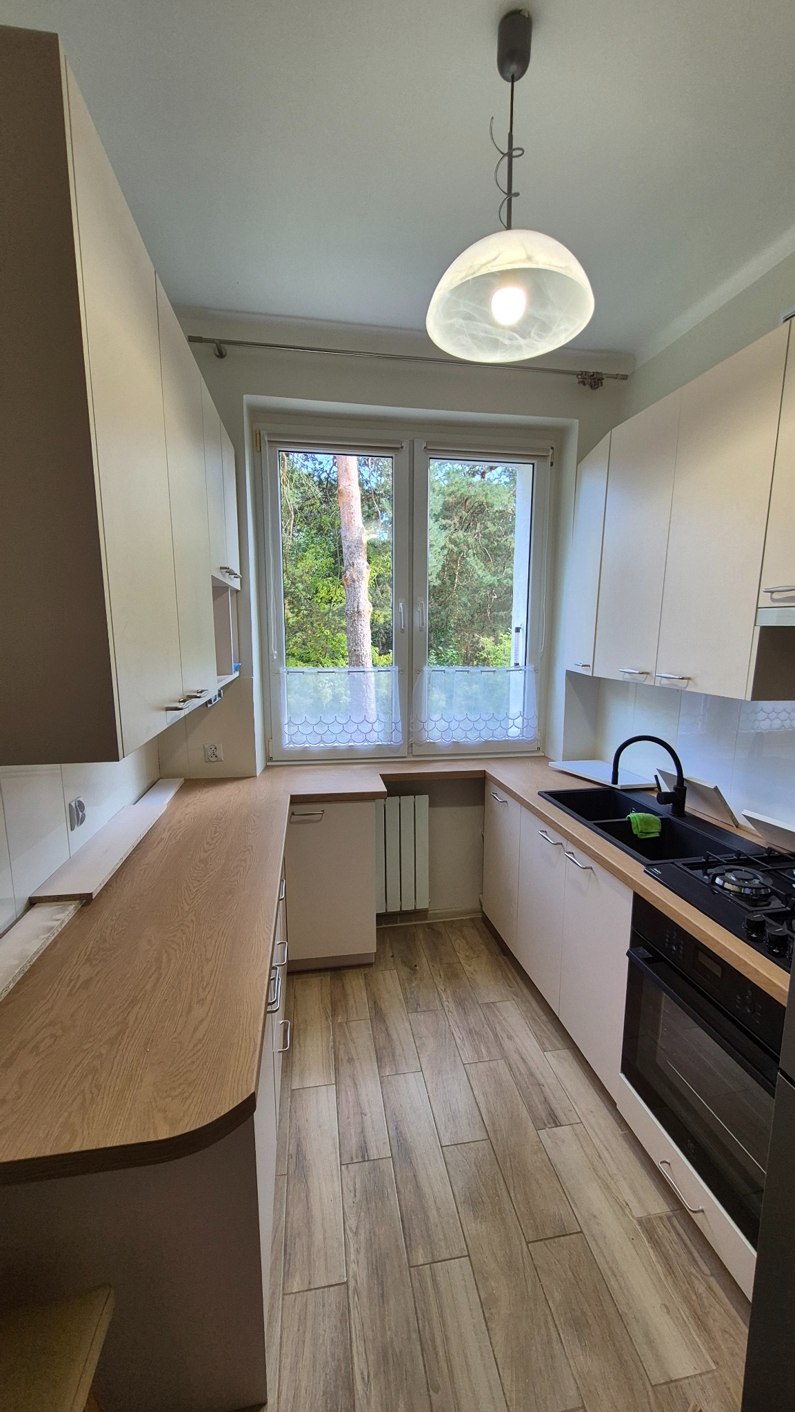 Jasna kuchnia z drewnianym blatem i widokiem na zieleń za oknem. Nowoczesne szafki, czarny zlew i płyta gazowa. Podłoga wyłożona płytkami imitującymi drewno.